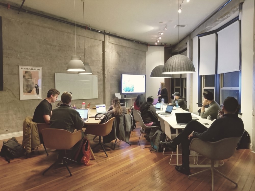 A group with laptops around a long table in a loft office, a presentation at the front